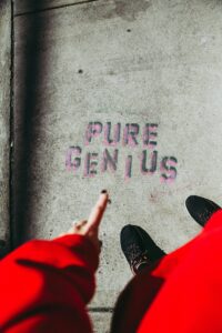 The words "Pure Genius" stamped on a sidewalk with a woman's hands and feet and bottom of red dress and pointing at it.